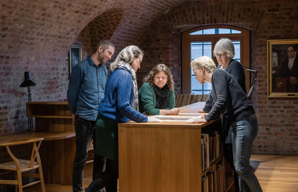 Fem mennesker som står rundt et bord og kikker ned på bilder