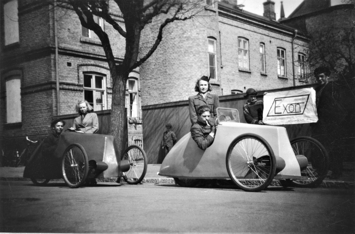 Reklamjippo för Exon Cykelbilsfabrik i Landskrona, enligt skylten de två pojkarna till höger håller upp. Två trehjuliga fordon med cykelhjul på gatan, troligen utanför ett av "Tvillinghusen". Uppgifter saknas. I "cykelbilarna" sitter en pojke och 
Tillhör samlingen med fotokopior från Hallands Nyheter som är från 1930-1940-talen.