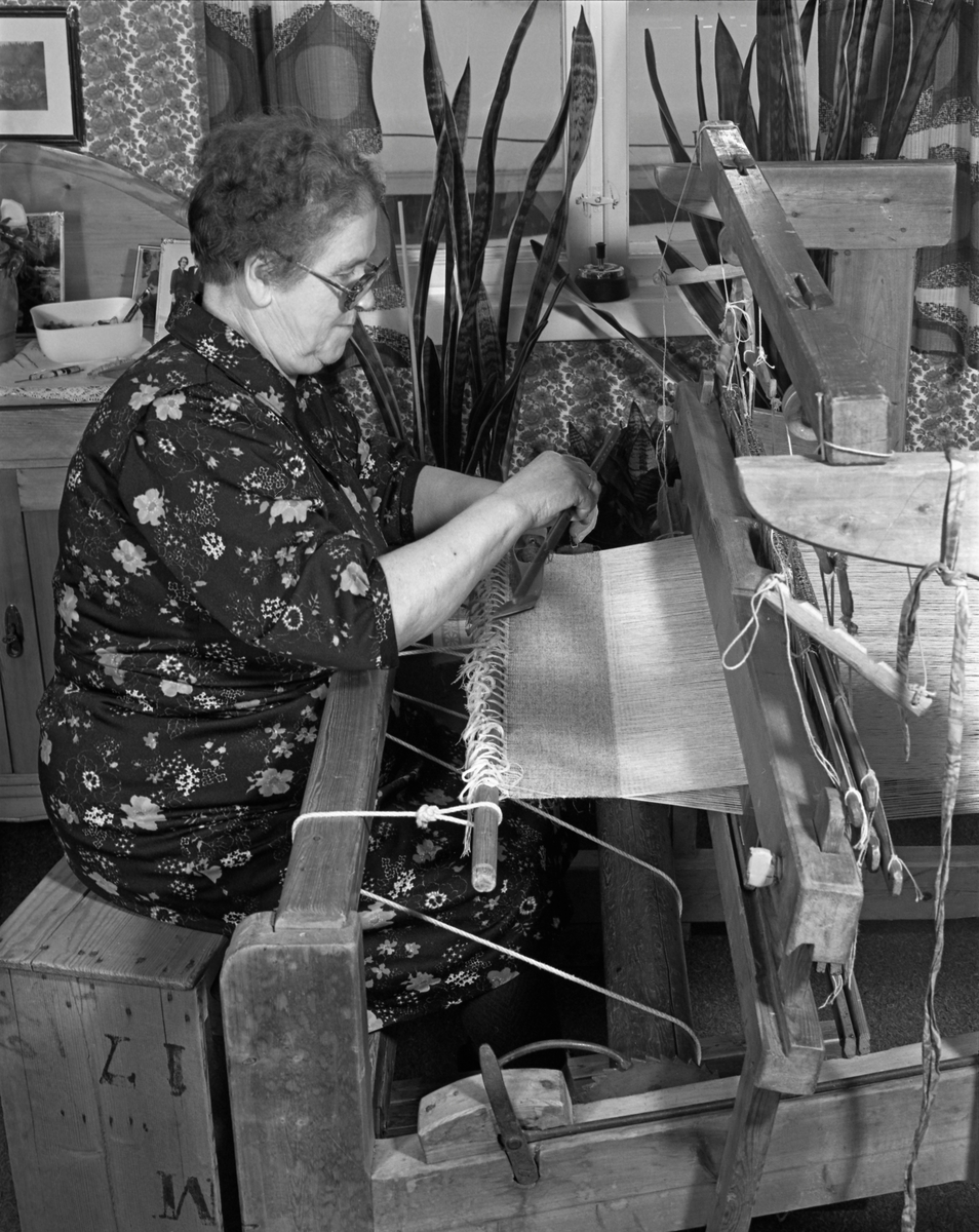 Nina Østerhaug (1916-1992) fra Nybergsund i Trysil, fotografert ved vevstolen i hjemmet sitt, der hun satt ved vevstolen og produserte stoff som hun seinere skulle sy busserull av. Nina var gift med [Paul] Henrik Østerhaug (1916-1990), som var tømmerkjører. Nina vevde og sydde mange av arbeidsklærne mannen brukte. Kontantsinntektene var beskjedne. Arbeidet ved vevstolen og bak symaskinen inngikk i strevet for å begrense pengebruken. Museet har flere fotografier av Henrik i arbeid, iført klær som Nina hadde produsert.