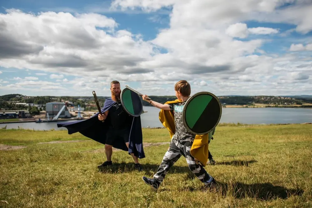 En voksen mann og en gutt ikledd middelalderdrakter kriger med sverd og skjold på toppen av et fjell