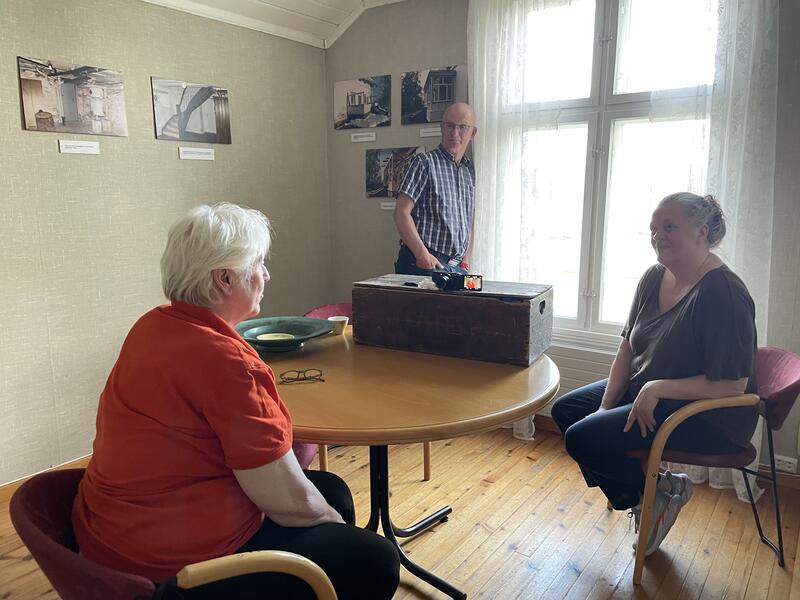 Foto av tre personer i et rom. To kvinner sitter ved et bord, den ene intervjuer den andre. Et kamera på bordet filmer hendelsen. En mann står i bakgrunnen.