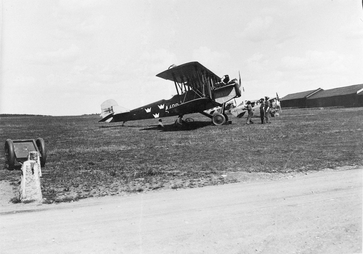 Arbete med uppställt flygplan B 1, Fiat BR, nr 4400, framför Flygkompaniets F-hangar på Malmen omkring 1924-1926. Sex personer vid flygplanet. Tre flygplan FVM Tummeliten i bakgrunden.