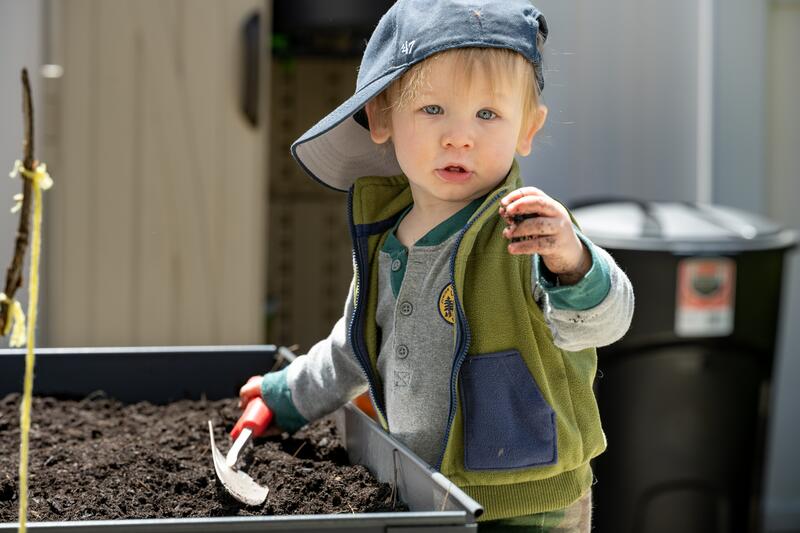 Liten gutt står ved en kasse med jord. Han viser frem et frø og har en spade i den andre hånda.