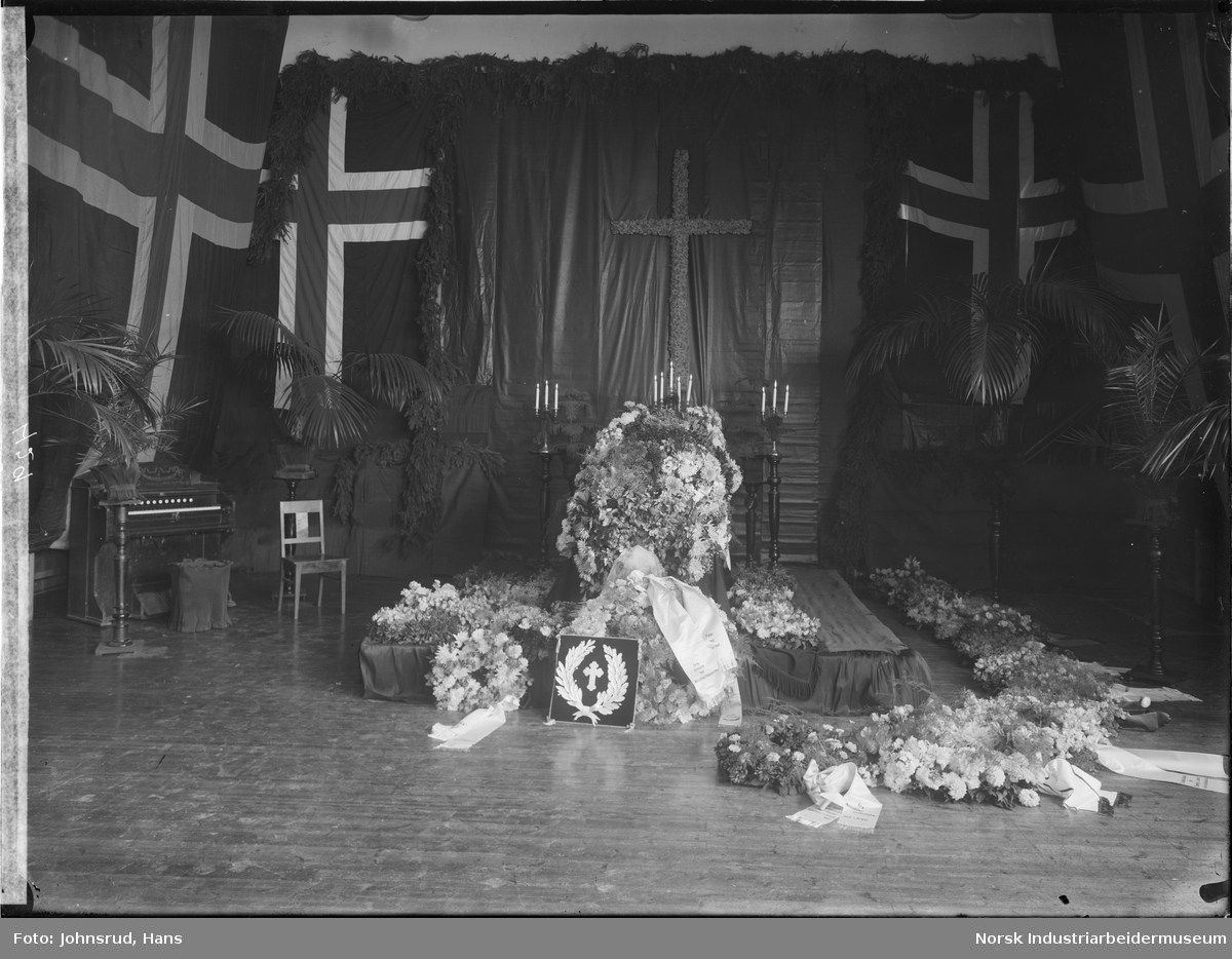 Begravelse til skolebestyrer Evje. Kiste omringet av blomster og blomsterkranser med tekstsløyfer. Norske flagg henger på veggene.