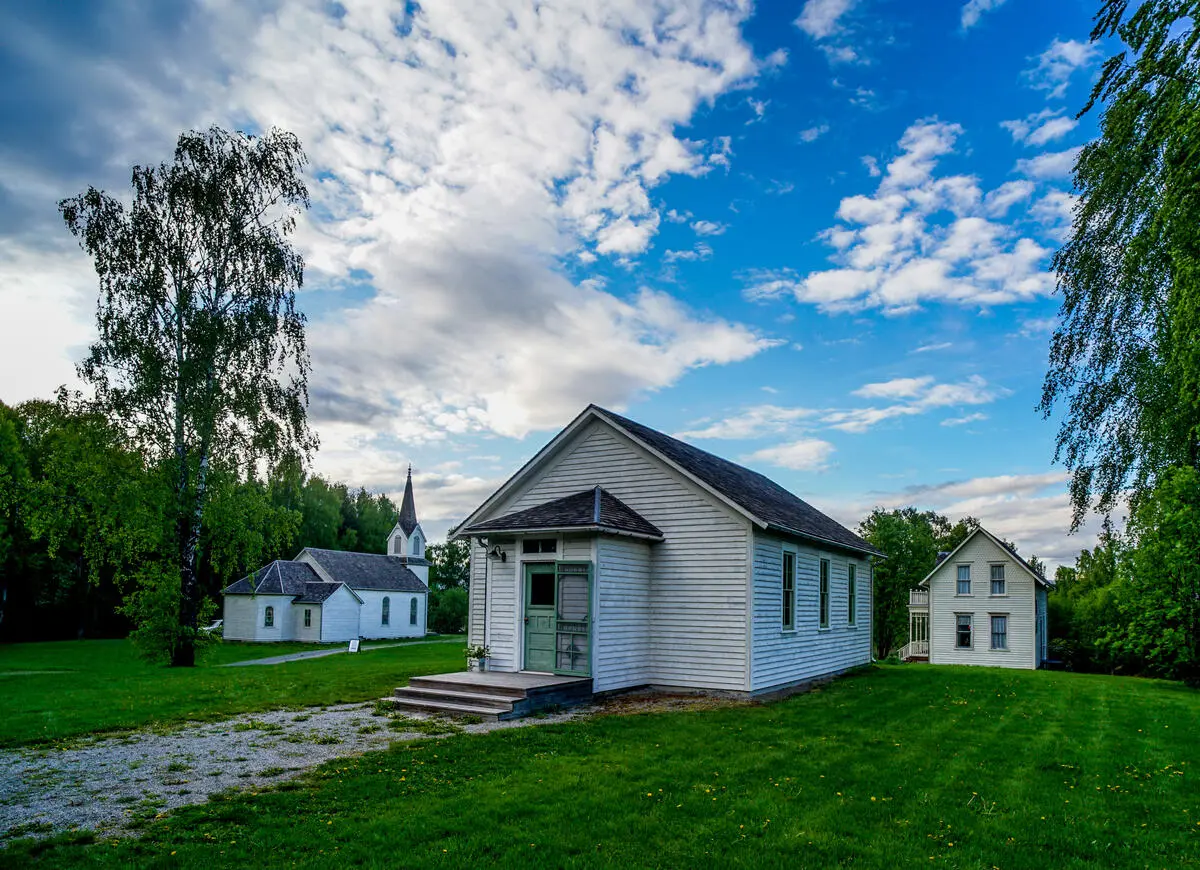 Et gammelt hvit hus med kobbergrønne dører står ute på et tun, i bakgrunnen står en gammel hvit kirke og et større gammelt hvitt hus