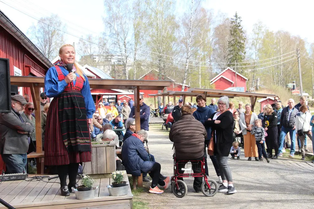 Foto av kvinne i bunad som står på et podium og holder tale. Mange mennesker i bakgrunnen.