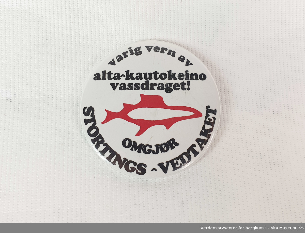 Rund hvit button i plast og metall, motivet er av en rød laks med teksten: "Varig vern av Alta-Kautokeinovassdraget! Omgjør Stortings-vedtaket".