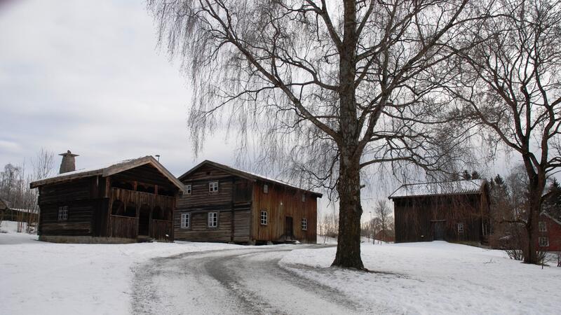 Tre 1700-talls tømmerhus står i et snødekket landskap med ei bjørk i forgrunnen.