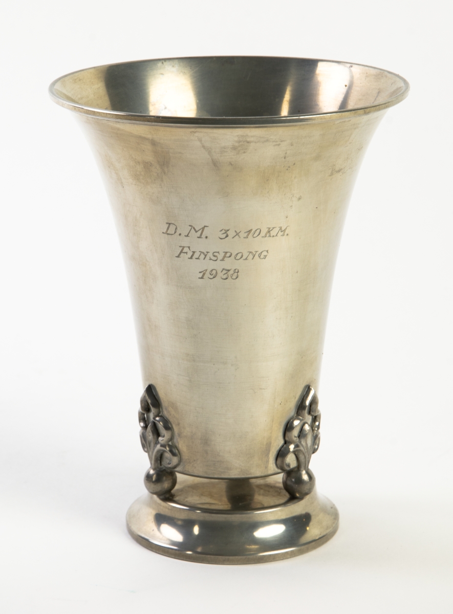 Pokal på tre kulfötter stående på sockel av tenn. Graverad med texten: "D.M. 3x10 km. Finspong 1938" 
Botten märkt: "L8, GAS, TENN, SVENSK VARA 24, 4, 2/17, 13".