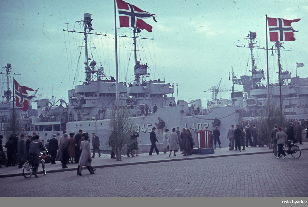 Folk spaserende på Rådhusplassen ved Honnørbrygga under feiringen av frigjøringsdagene 1945, trolig 13.mai. ..Fartøyene på bildet er havgående minesveiperene tilhørende RN 40th Minesweeper Flotilla som ledet styrken inn Oslofjorden. Nærmest HMS Cynthia J345 og HMS Elfreda J402.