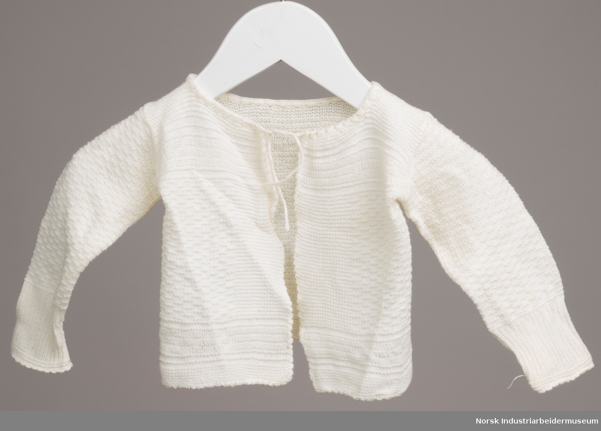 Hvit trøye til spedbarn med snor i halsen. Trøyen er maskinstrikket med mønsterstrikk i samme farge. Masker er løse i venstre armhule.