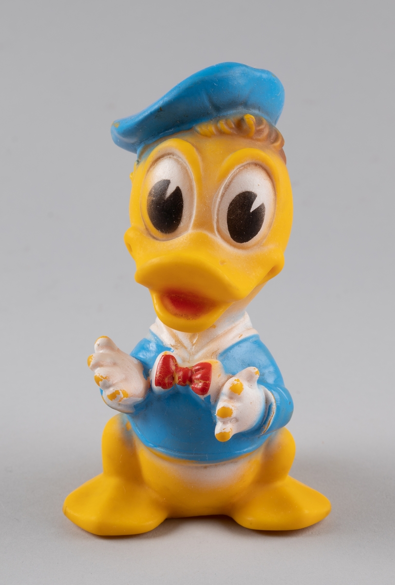 Leketøy av mykplast i form av en Donald Duck. Den er gul med blå jakke med rød sløyfe, den har blå hatt og hvite hansker. Øynene er store og ovale, hvite og svarte.