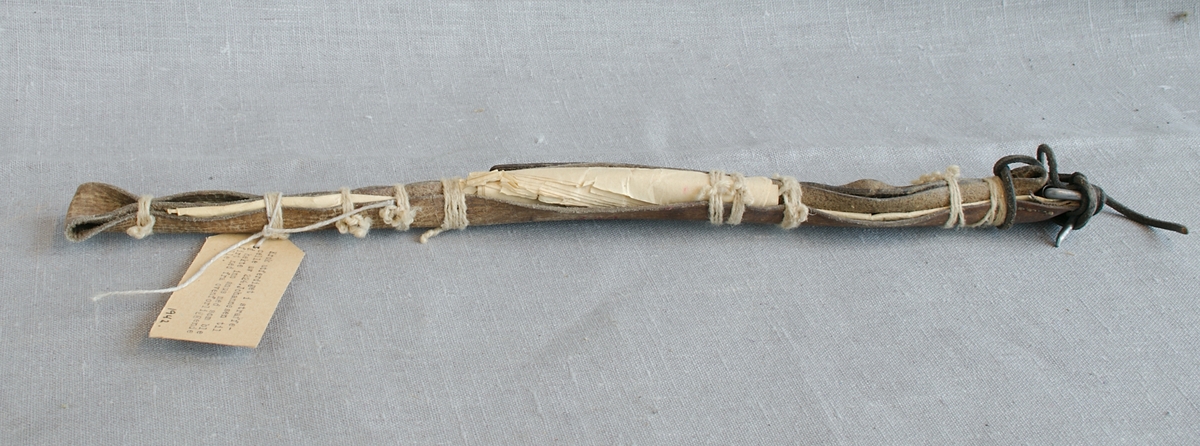 Krok laget av metall med et langt skaft laget av en lærreim, trolig et belte. Oppstivet med sammenbrettet papir og surret med hyssing.