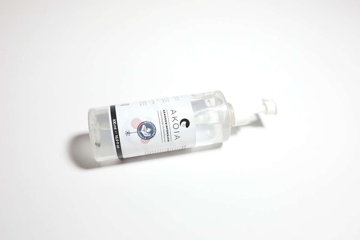 Sylinderformet flaske som smalner inn på toppen mot en pumpetut. Rundt midten av flasken er det festet en etikett med informasjon på norsk.