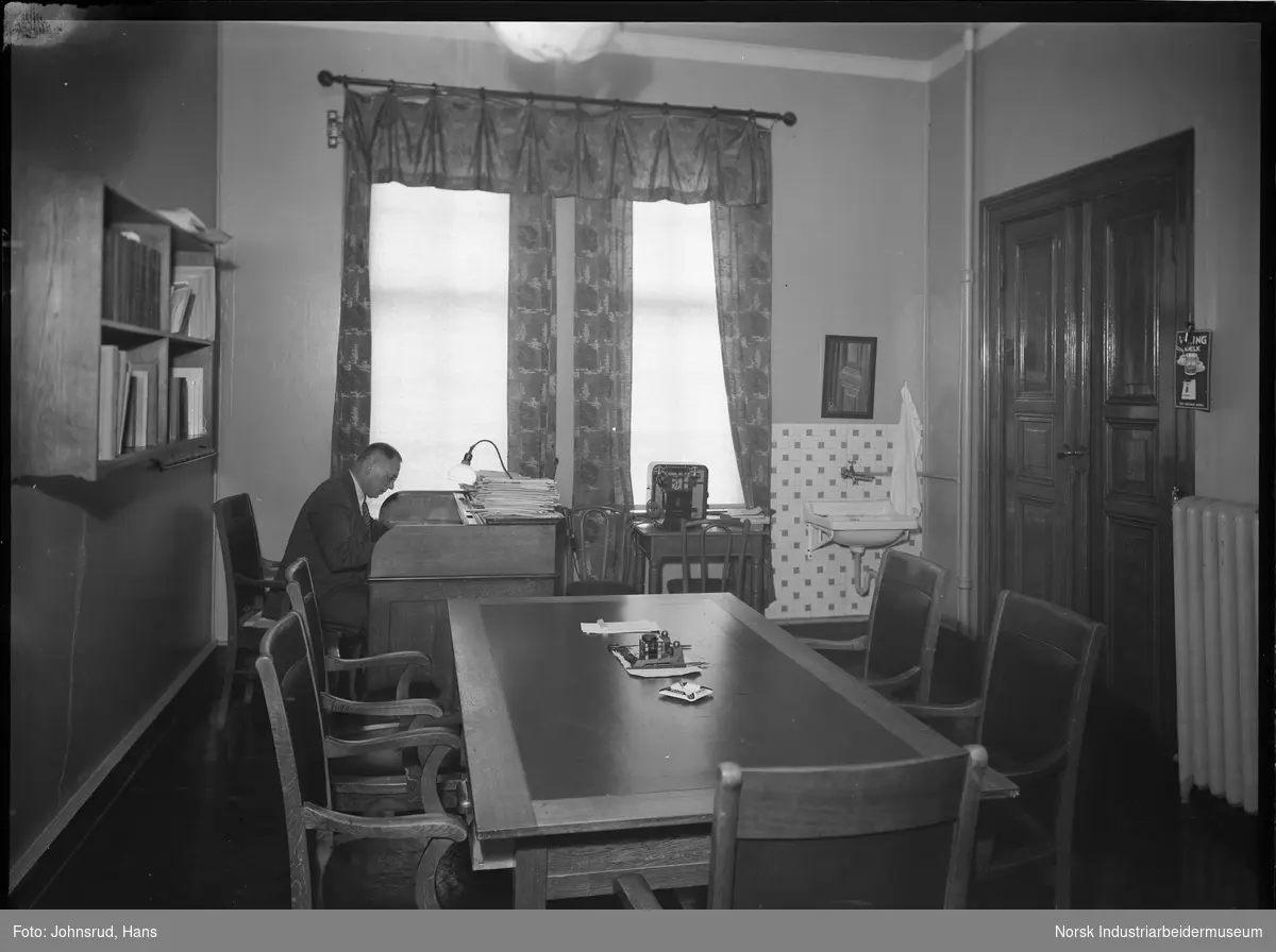 Kontorlokale til Kongsberg Sparebank. Mann i arbeid bak kontorpult. Telefon stående på bord ved siden av, vask på veggen bak inngangsdør til kontoret.