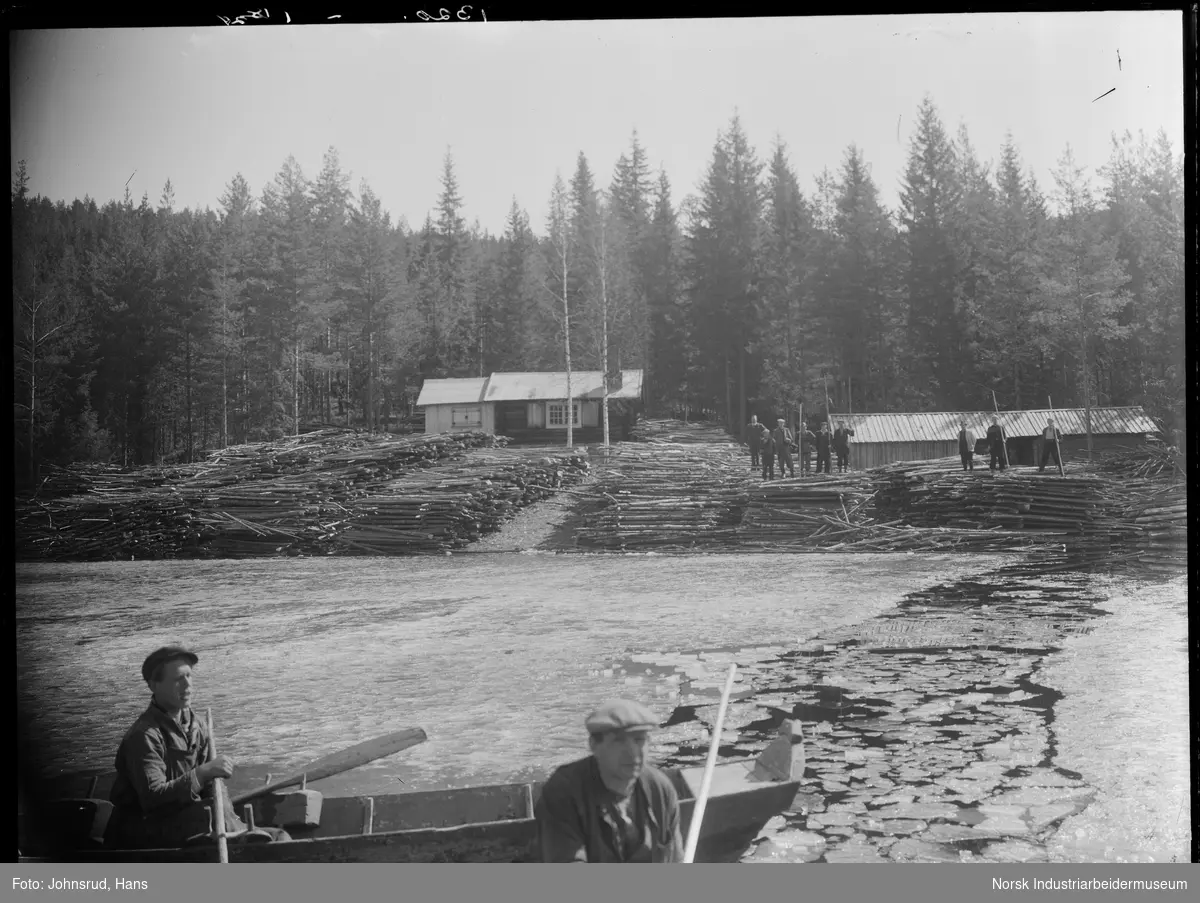Vedlager ved Elgsjø. Tømmer ligger i stabler langs land nedenfor hytte. Menn i robåt i islagt innsjø. Menn med fløtestokker stående på tømmerhaugene på land.
