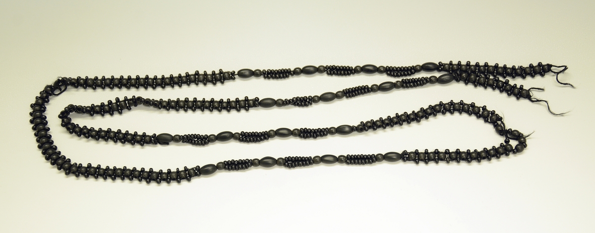 Fra protokoll: Halskjede. Større og mindre runde og ovale perler i sort matt. Knyttet i skiftende mønstre. Låsen mangler.
