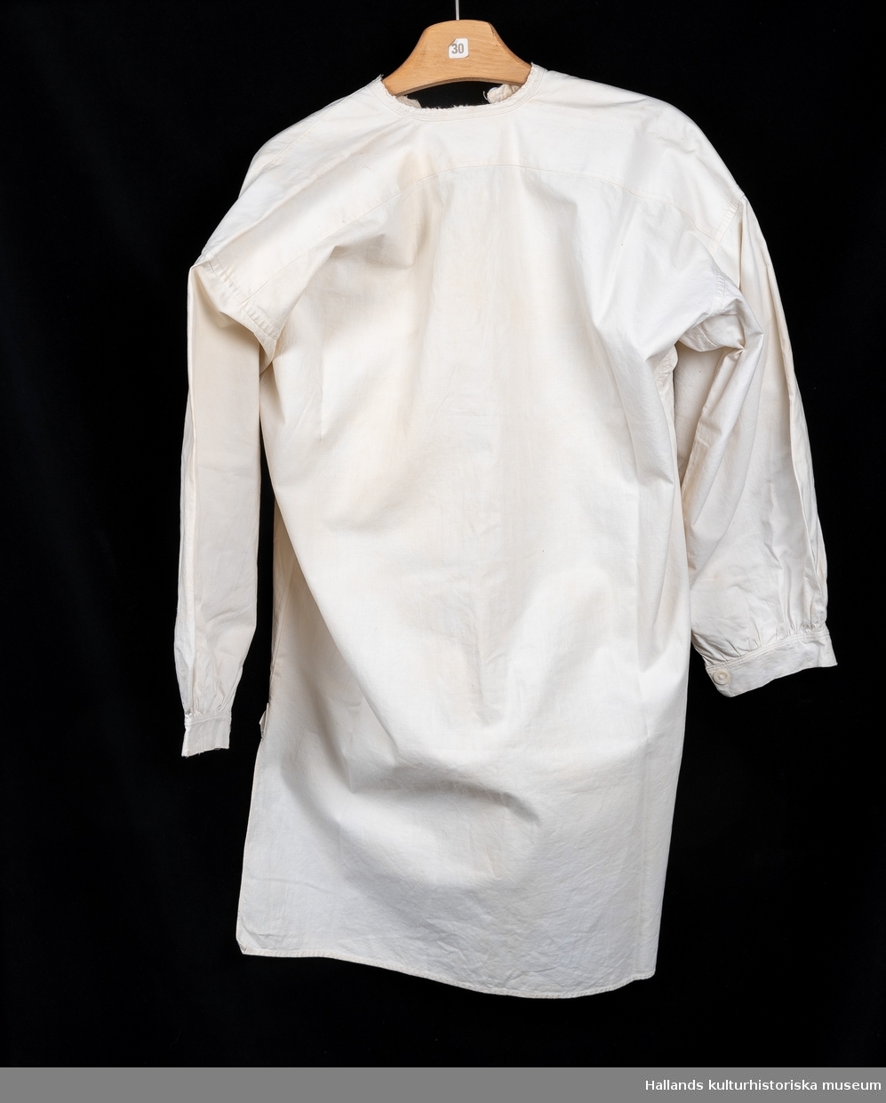 Skjorta, så kallad slåtterskjorta, använd vid slåtter. Av vit bomull. Bakstycket går något längre ner än framstycket. Långärmad med knapp vid manschetten. Sprund med knapp vid halsen. Rund hals. Märkt: "K", med korsstygn. En lagning. Lite sliten i kanterna.