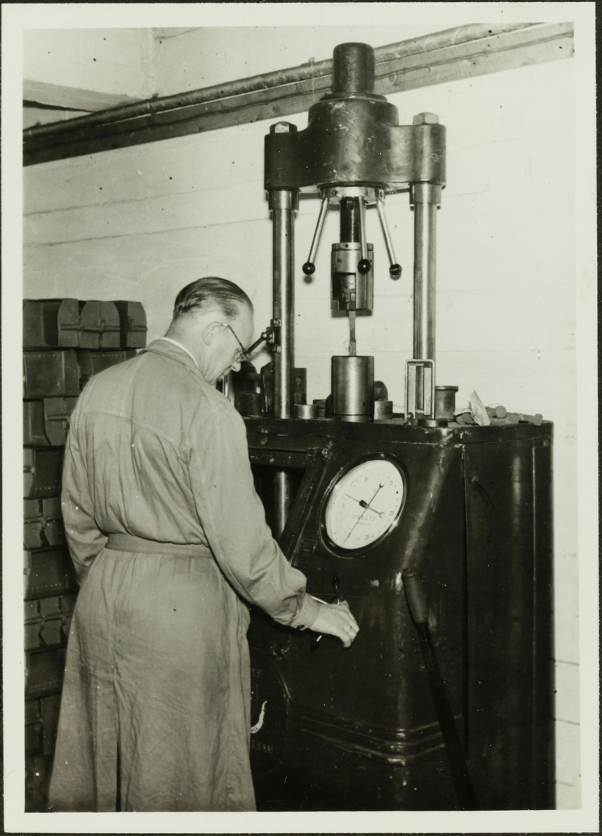 Ein mann i frakk står framfor eit stort apparat i eit rom (laboratoriet?) på Sandnes Auducerverk.