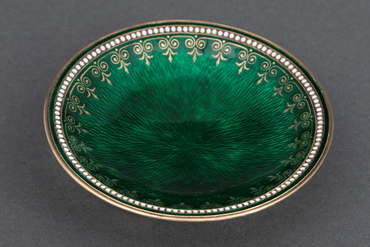 Lav, rund skål i forgylt sølv med tre kuleformede ben. Innvendig er skålen dekket av grønn, halvtransparent emalje over håndgravert mønster. Langs kanten er det en forgylt palmettbord og en perlerad i hvit emalje.