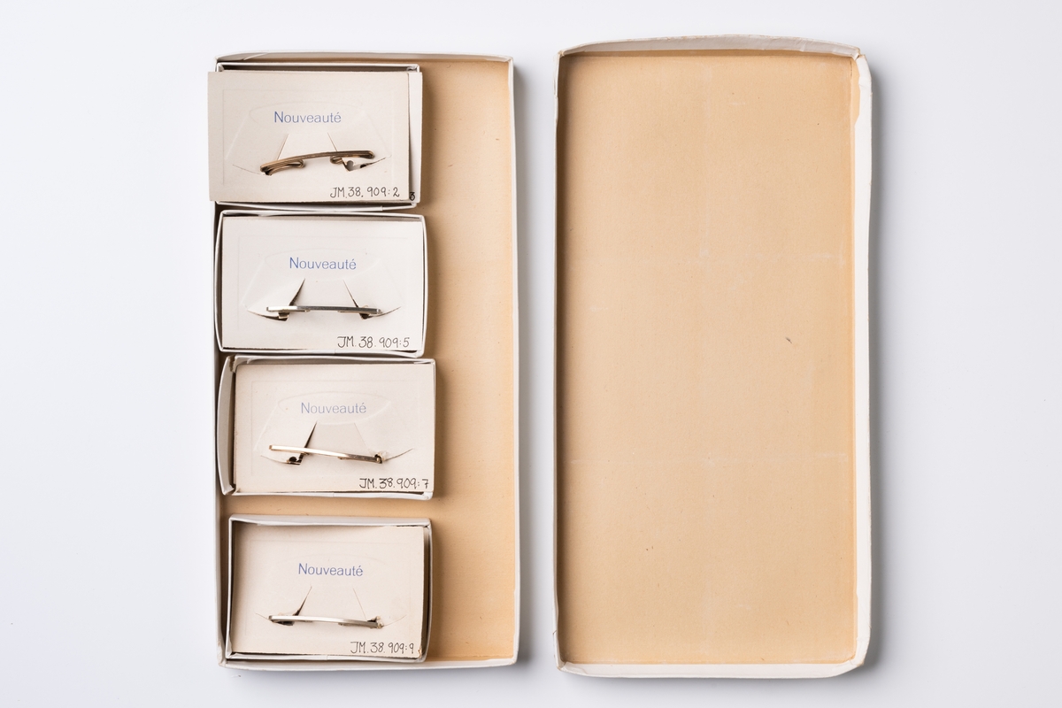 Kartong för kraghållare av märket Nouveauté, 9 st. Förpackningen är av papper och består av låda och lock. Kraghållarna sitter fast på pappbitar med tryckt text: "Nouveauté".