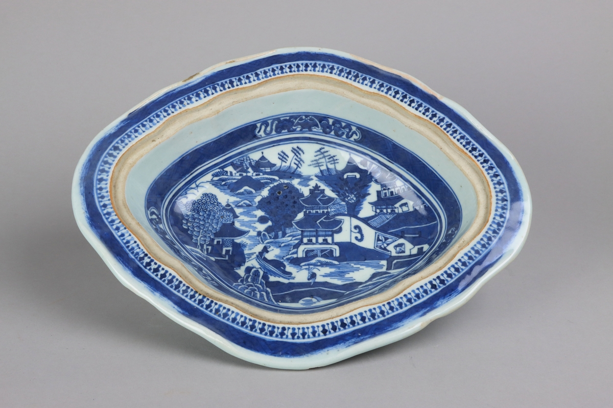 Oval broken form with a wide flat rim, decorated with a dark blue border with criss cross pattern. The inside of the dish slightly everted and undecorated. The well surrounded with a border of a criss cross pattern and rectangular reserves filled with symbols of good fortune. Centered in the well landscapes scenes with pagodas, buildings, waters, bridges and gardens. The outside of the dish is decorated with rose branches All decor in blue underglaze. The base of the dish without glazing.