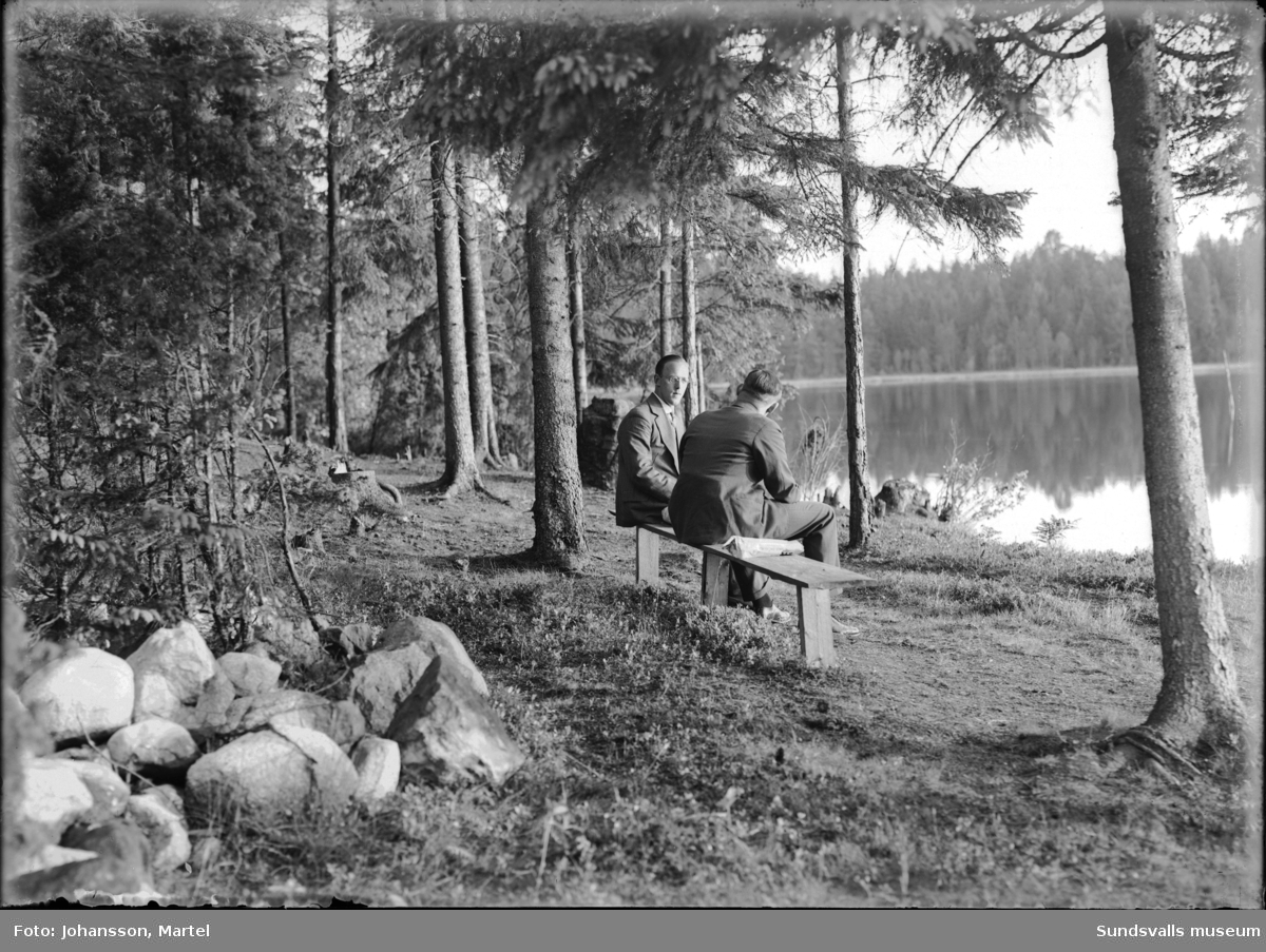 Gruppbild i det gröna med män, kvinnor och barn. Tvåa från vänster är fotografen själv, Martel Johansson. På den andra bilden sitter två av männen på en bänk invid en skogssjö.