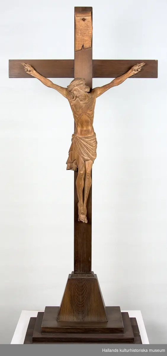 Skulptur av trä. Plattform och kors av mörkbetsat trä (furu). Jesusfigur handsnidad av ädelträ (troligtvis valnöt). Ovanför Jesusfiguren en skylt: "INRI". Svårläst signatur i blyerts på figurens rygg: "NJ??? 15/11 29".