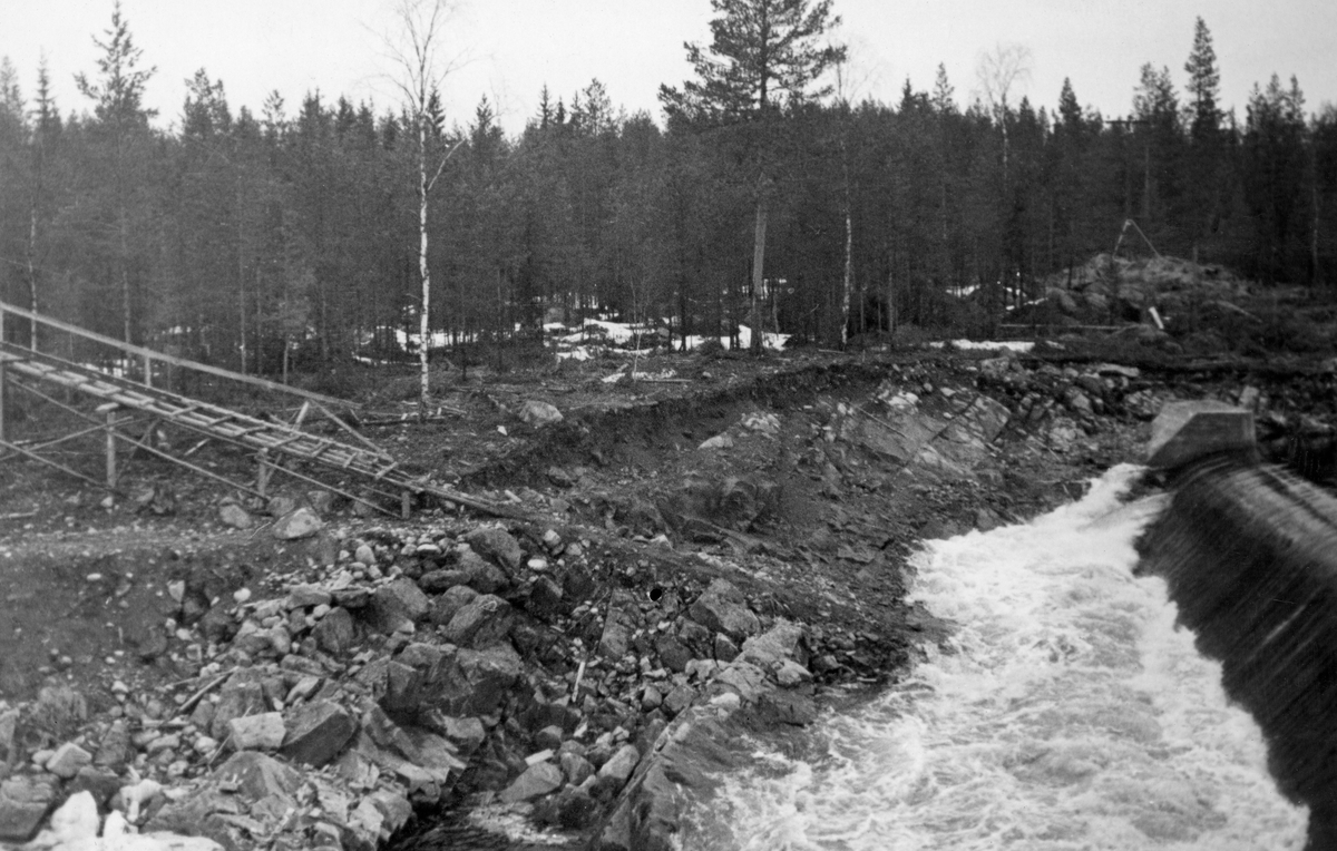 Dammen som skulle stuve opp vann til Kvernfallet kraftverk i elva Søndre Osa i Åmot kommune i Hedmark, fotografert våren 1936, like etter at anleggsarbeidet var ferdig. Her renner vannet over en avrundet terskel, utført i armert betong. Denne konstruksjonen er plassert på tvers av elveløpet, like framfor ei oppoverskrånende bergflate, som leder vannet fra nedfallssona mot den søndre elvebredden, før det føres videre vestover i det gamle elveløpet. Turbinrøret, som skulle lede vann nedover mot kraftstasjonen ved Brufallet, var også plassert på sørsida, litt til høyre for dette bildeutsnittet. Til venstre i bildet ser vi et trestillas som ble reist i forbindelse med anleggsarbeidet.

Litt mer informasjon om kraftutbygginga i Søndre Osa, og om anlegget ved Kvernfallet spesielt, finnes under fanen «Opplysninger».