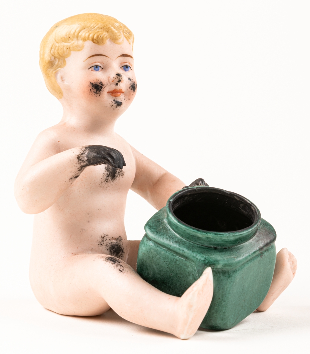 Bläckhorn i form av en naken pojke som sitten med en grön burk framför sig. Pojken är blond med svarata händer och bläckfläckar i ansiktet. Ihålig, av keramik.
