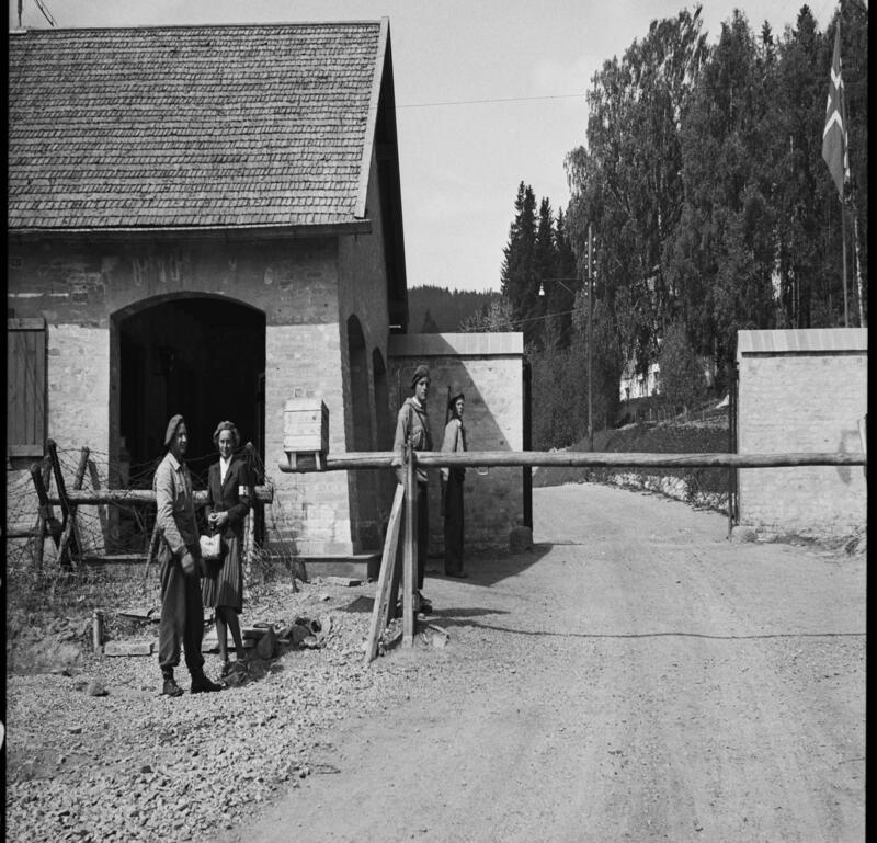 Fotografi av ytre port. To lave murvegger med vei mellom, foran er det laget en sperre av tømmer. Fire mennesker står til venstre i bildet.