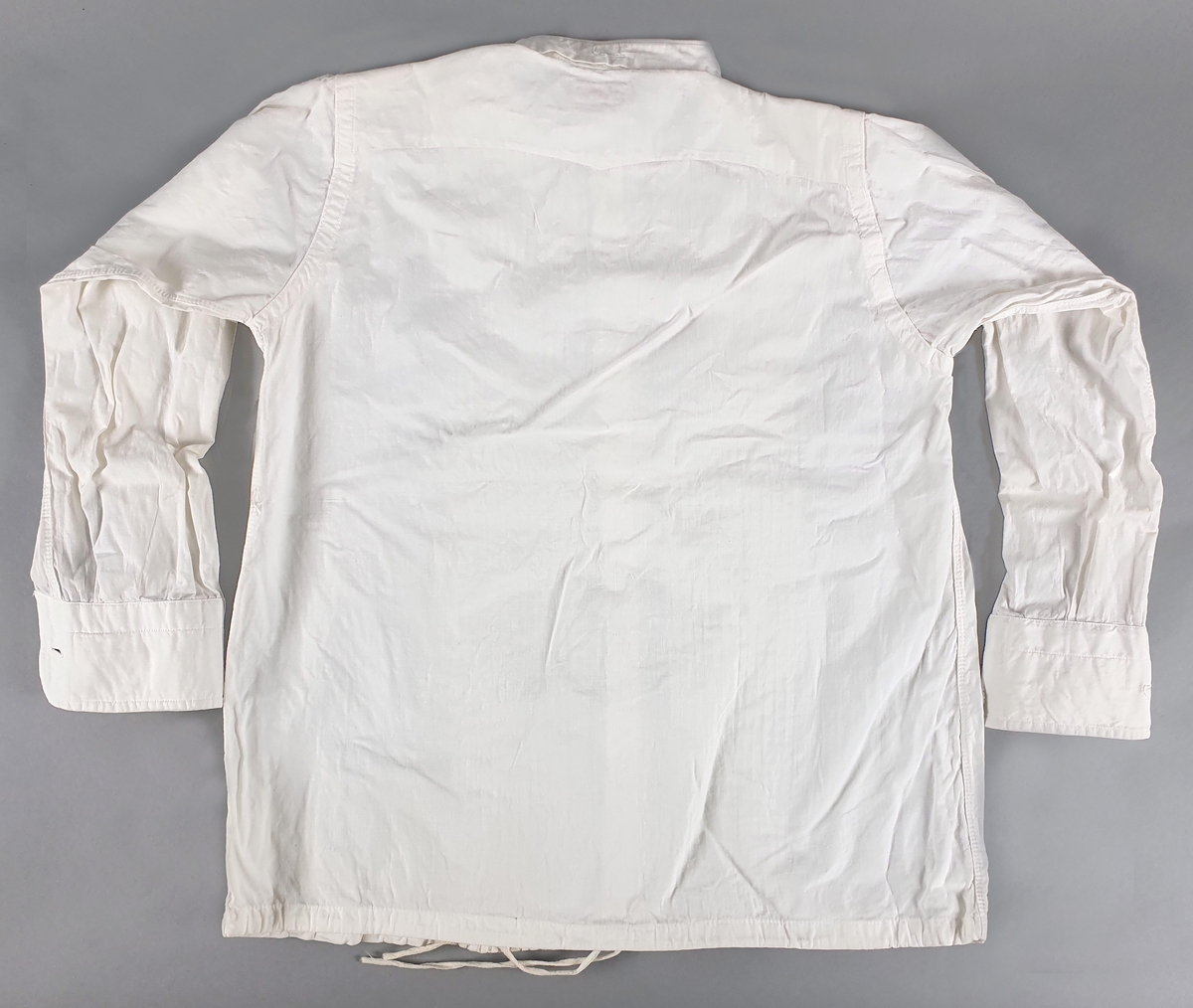 Skjorte av bomull, med vertikale dekorfolder på brystet, snor nederst. Smal linning i halsen med svart mansjettknapp. Hvite knapper midt foran. Skjortene er stivet.
