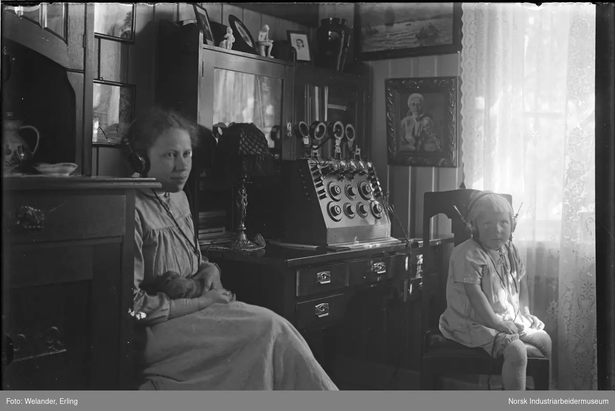 Kvinne og barn sittende i stue med øreklokker på hodet. Det lyttes sannsynligvis på en radio som står på ett bord mellom dem. Kvinnen har en valp i hendene.