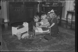 Tre barn leker på gulv i stue. En av jentene sitter med en b