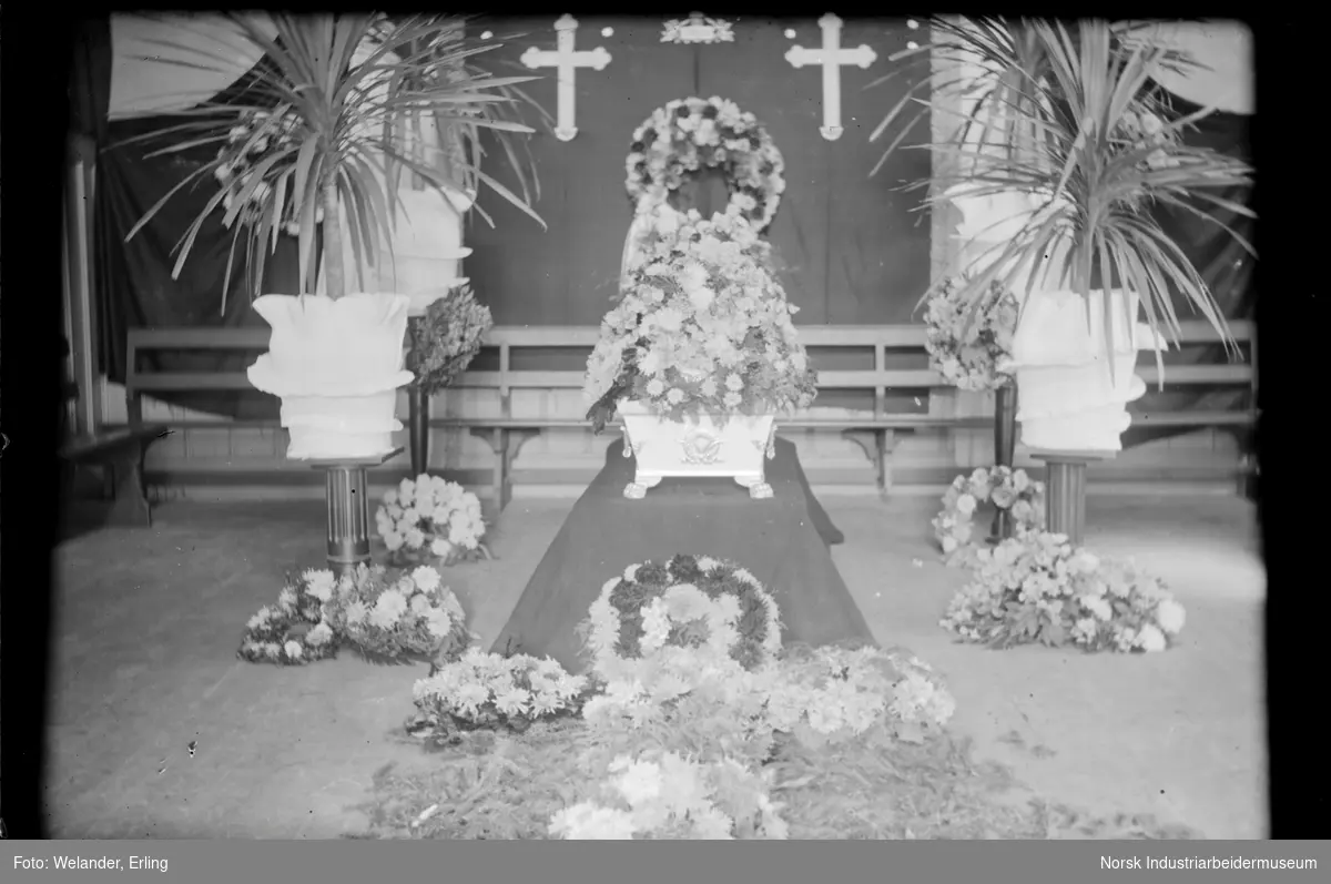 Begravelse i Metodistkirken (Rallarhuset) på Rjukan. Kiste stående midt i rommet dekket med blomster. Blomsterkranser liggende rundt på gulvet. Potter med palmer på hver side av kisten.