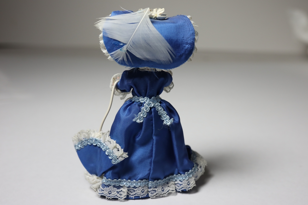 Dukke fra Belgia, stående, med blå kjole i kunstsilke med hvite blondekanter, liknende hatt og parasoll. Har et forkle rundt livet med tekst.
