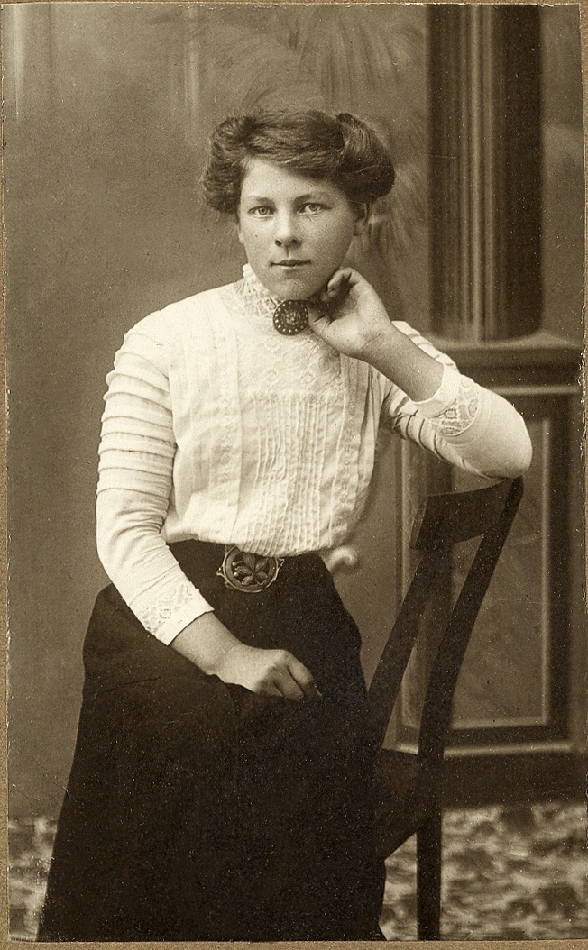 En kvinna i ljus höghalsad kjol och mörk kjol sitter på en stol. 
Under fotot text med blyerts: "Helga".
Knäbild, halvprofil. Ateljéfoto.