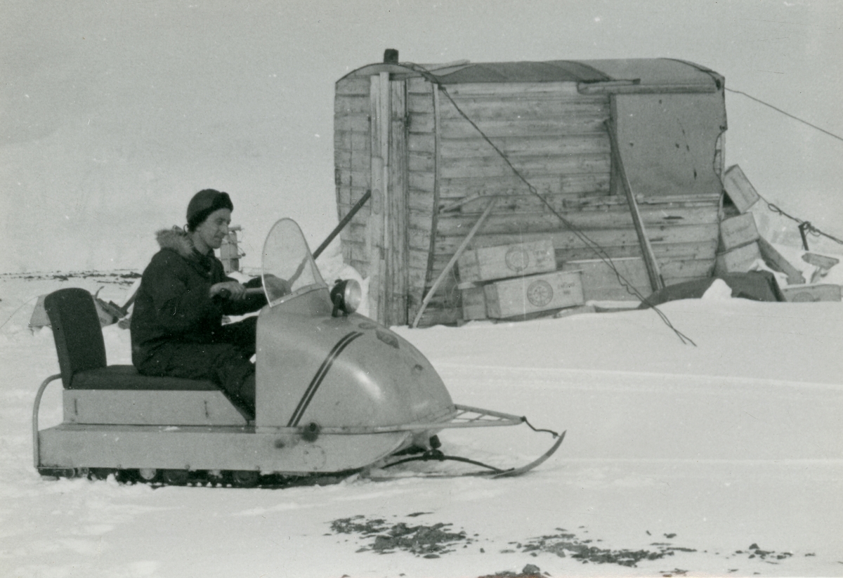 Mann på snøskuter foran en liten hytte på begynnelsen av 60-tallet