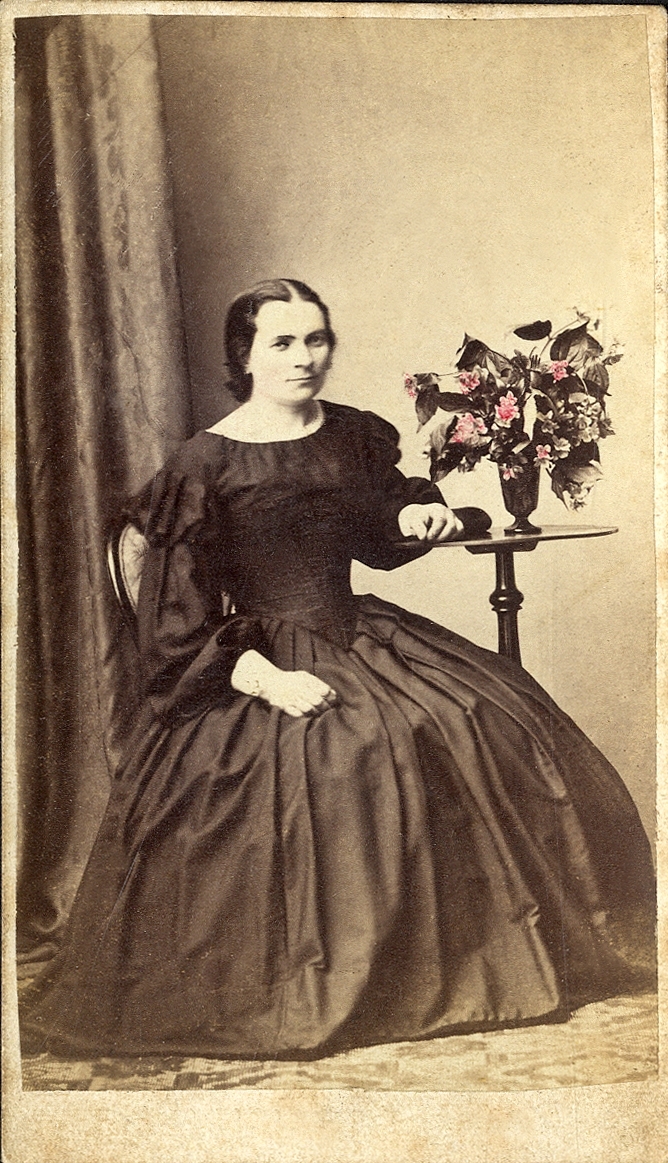 En kvinna i mörk krinolin sitter vid ett pelarbord med en blomvas på. 
Blommorna är delvis handkolorerade. 
Helfigur. Ateljéfoto.