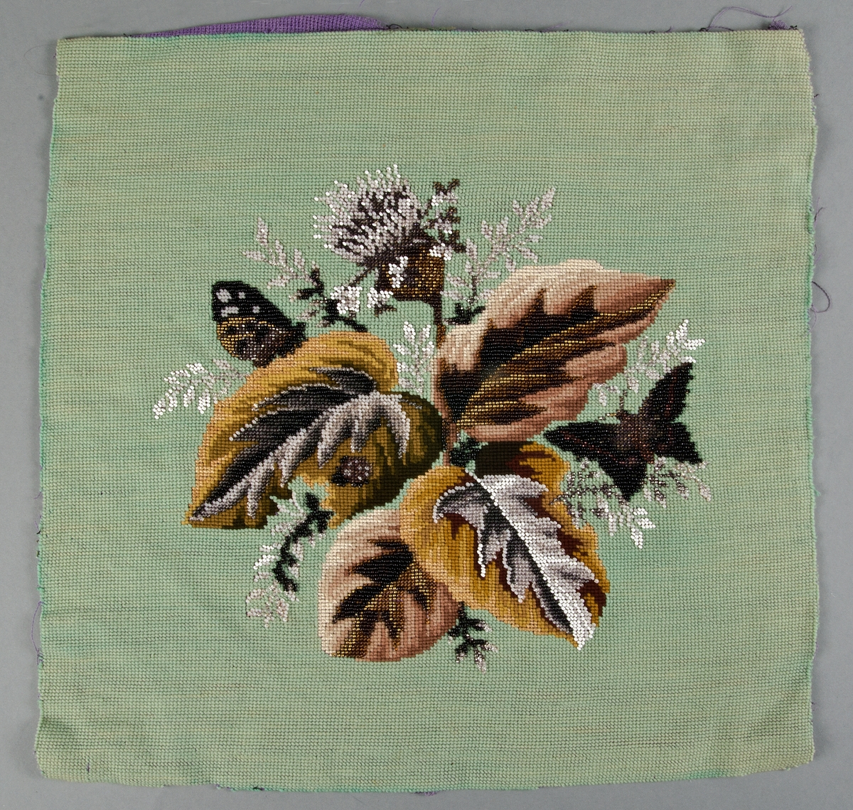 Korsstygnsbroderi i ullgarn, silke och pärlbroderi på lila stramalj. Vitt, svart, grått, gult och brunt samt mässings- och stålpärlor. Föreställande blad, blommor, och en fjäril.