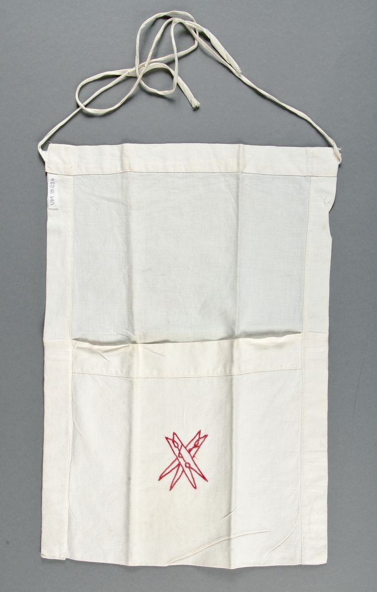 Klädnypspåse av vit bomullslärft i form av förkläde med stor ficka. Knytband. Två klädnypor broderade i rött bomullsgarn i stjälksöm.