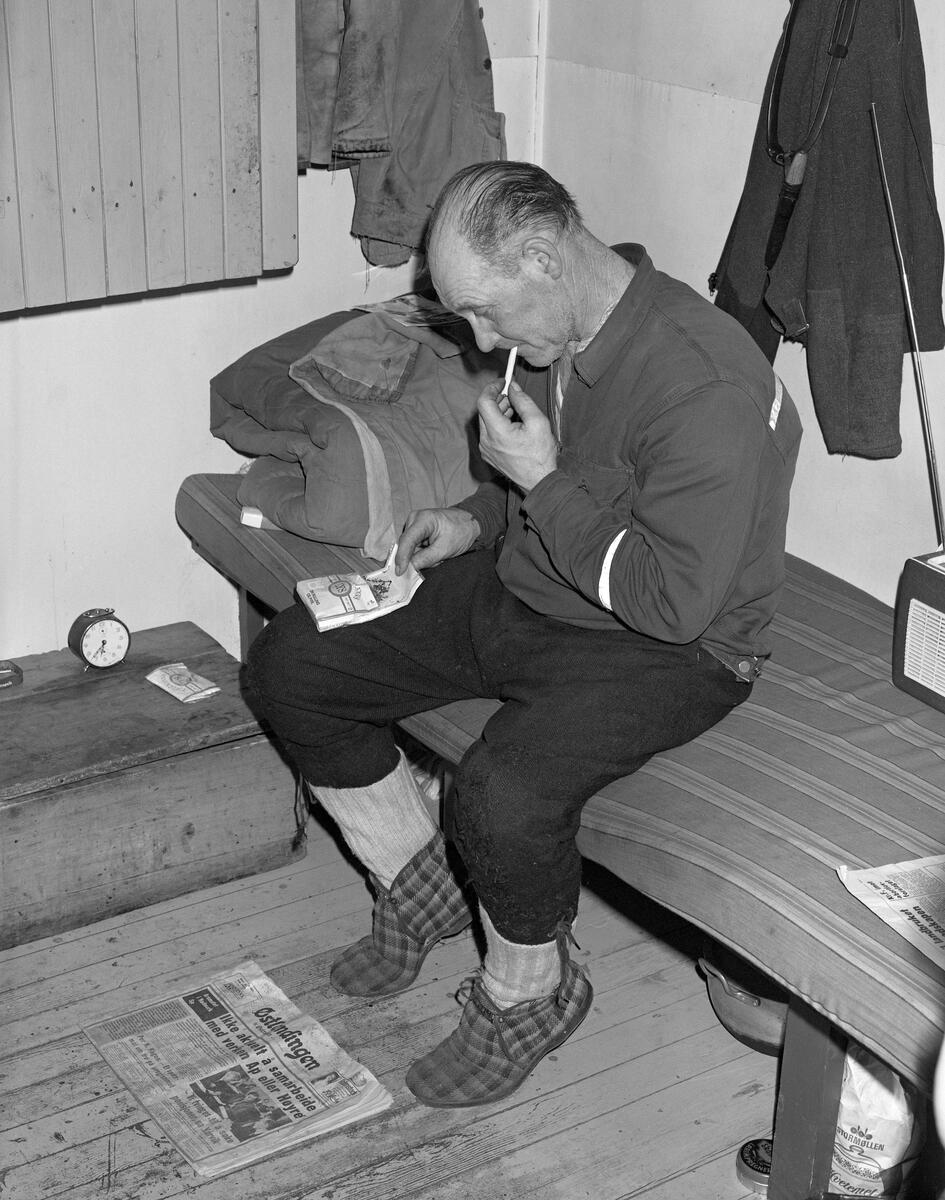 Skogsarbeider hviler i koia. Johan Rasch ruller seg en sigarett mens han sitter i senga si i Smaltjernkoia i Svartholtet, Elverum, Hedmark i januar 1975.