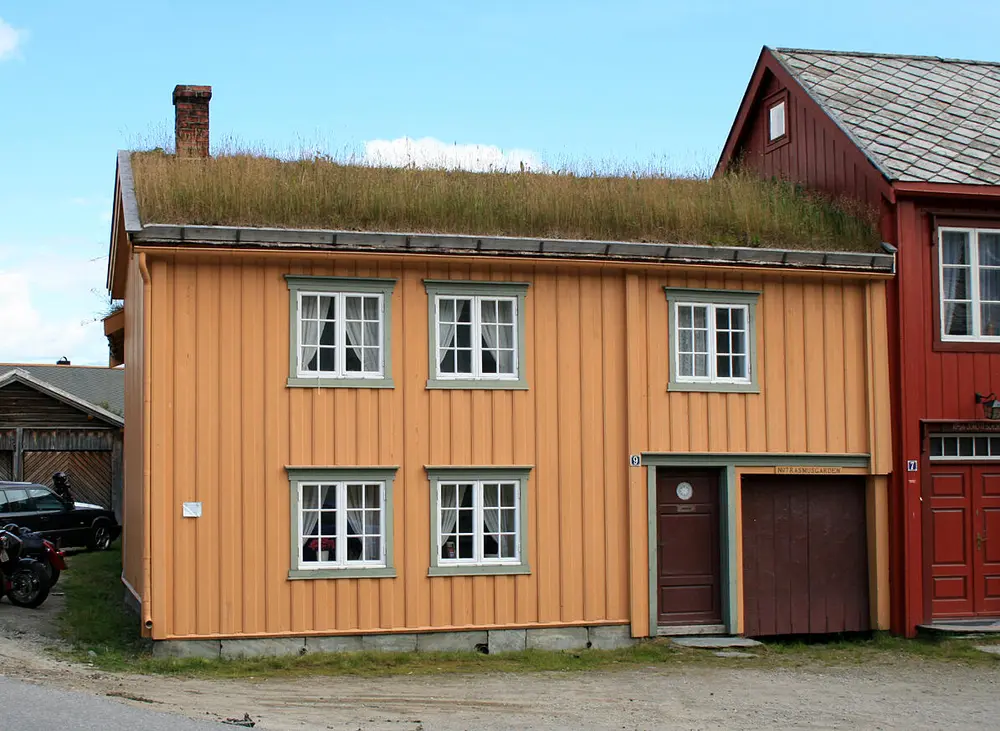 Bildet viser et gult trehus med gresstak. Det er to etasjer, ei inngangsdør og en port. Man ser at bygget ligger i ei gate med bygg på begge sider.