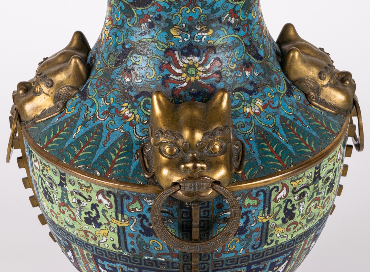 Urna. Metall. Kinesiserande stil. Fyra mässingshandtag i form av demoner med ring i munnen. Tandlist i relief. Blå botten med rik dekor i polykroma färger.