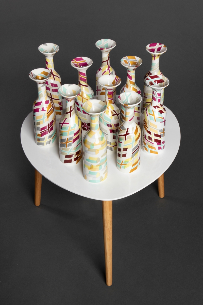 11 porselensvaser på et hvitt bord med tre trebein. Vasene er dekorkert med dekaler som viser variasjoner over det samme flagget. En av vasene har hus som motiv.