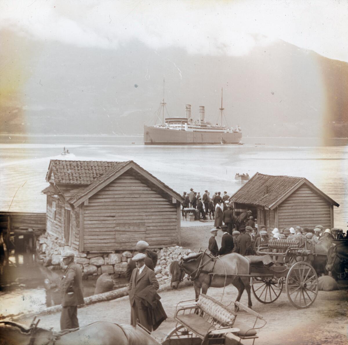 Dias. Passasjerskipet "Stavangerfjord" ligger på fjorden ved Loen. I forgrunnen står flere grupper med kvinner og menn på en brygge og blant uthus ved sjøen.  