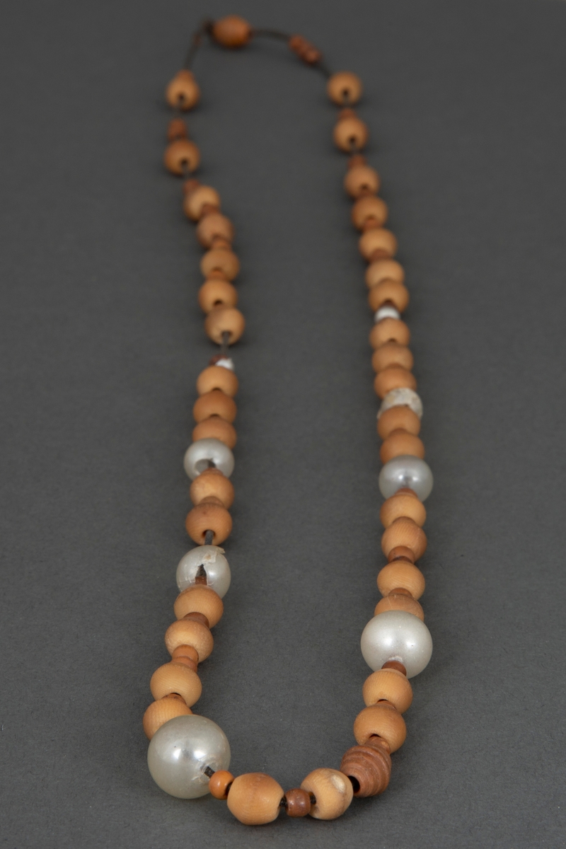 Perlehalssmykke bestående av større og mindre, dreide benperler, lysebrune og brune, og noen hvite glassperler. Perlene er tredd på en svart, noe elastisk tråd som har strukket seg sterkt.