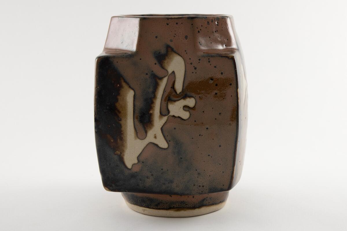 Vase med fire vegger og åttekantet munning.  Bygd opp i steingods glasert med persimonglasur (brun m. svarte spetter), og  penseldekor lagd med voksresist.  Dekor på alle fire sidene av vasen.