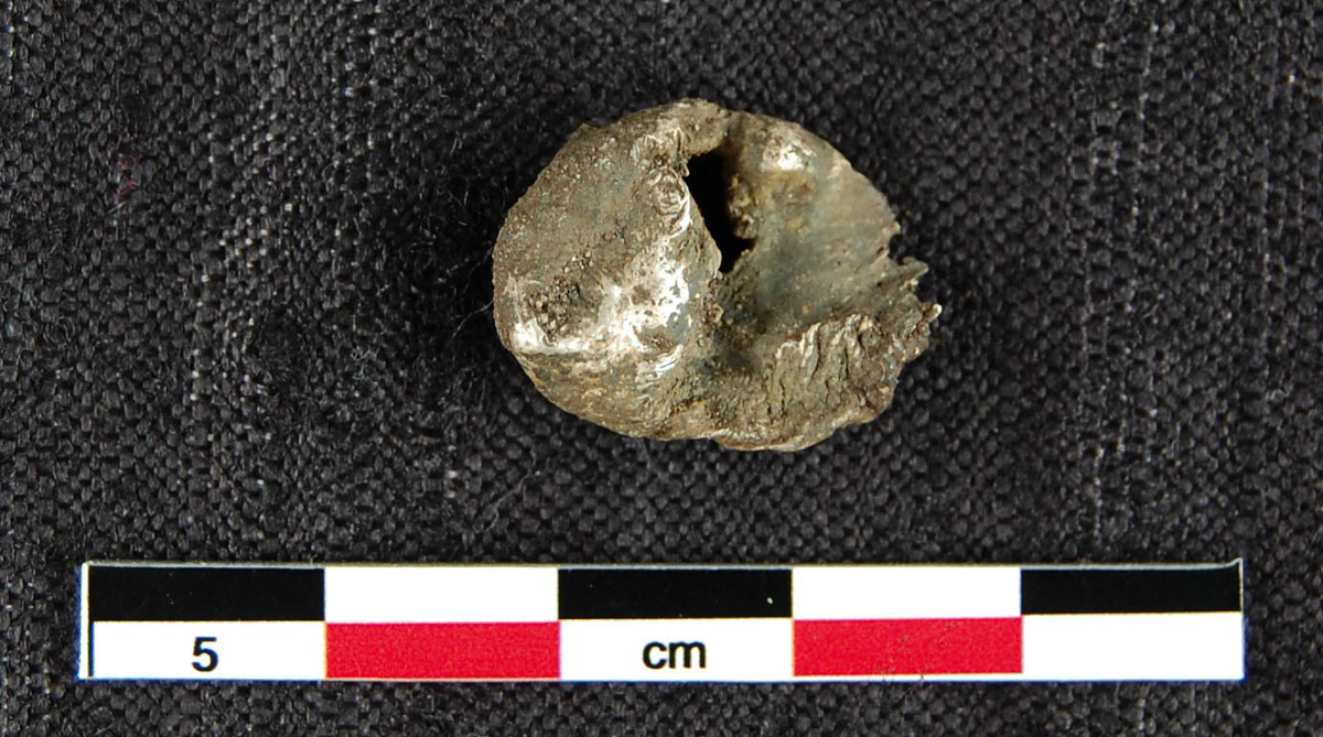 Oval, irregulær klump av smeltet sølv, med et rektangulært hull i. Mål: T: 0,74 cm. Stl: 1,86 cm. Stb: 1,45 cm. Vekt 4,49 g.