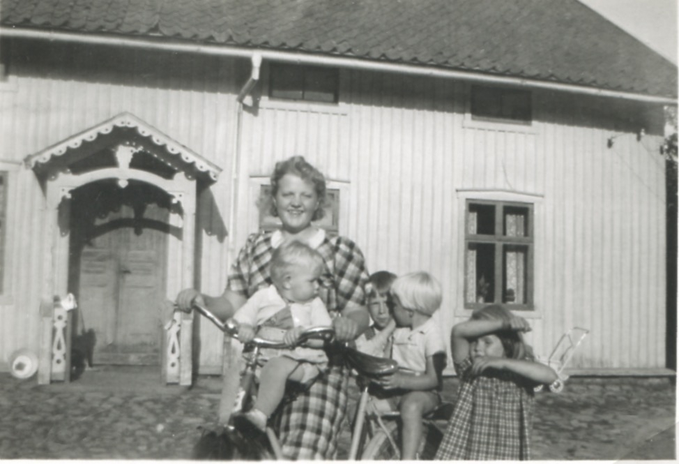 Kerstin Wessberg (1925 - 1990) på cykel tillsammans med fyra små barn framför gården Eken 1:1, okänt årtal. Kerstin var dotter till Amos och Hildur Wessberg. Hildur var dotter till Alm, tidigare ägare av gården Eken.
